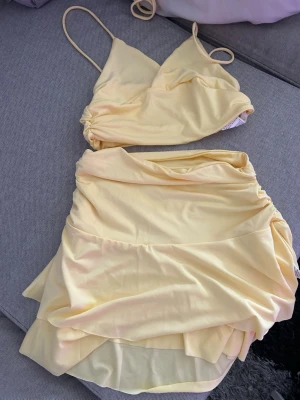 Gult tvådelat set med omlottkjol - Snyggt tvådelat set i ljusgul färg med crop top och omlottkjol. Toppen har smala axelband och lätt rynkade detaljer, kjolen är draperad och har lager på lager-effekt. Mjukt och stretchigt material, perfekt för en trendig look.