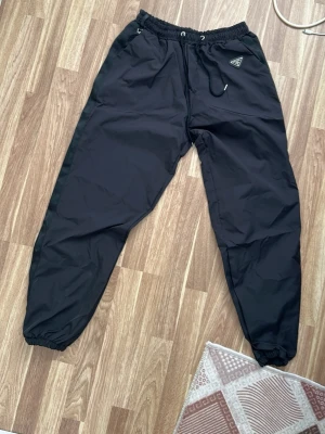 Svarta  från Prada - Svarta byxor från Prada med elastisk midja och dragsko. Byxorna har resår vid bensluten och en liten trekantig logotyp på vänster sida. Tillverkade i ett slätt, sportigt material som känns lätt och bekvämt.