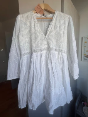 Vit broderad kortklänning med spets - Superfin vit kortklänning från Zara med broderade detaljer, spetskant och v-ringning. Klänningen har långa ärmar, knappar framtill och är tillverkad i luftig bomull. Perfekt för sommarens alla tillfällen! Köpt från sellpy med lappen kvar!