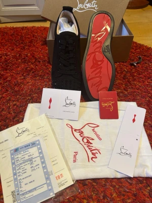Christian Louboutin svarta sneakers - Svarta sneakers från Christian Louboutin med klassisk röd sula och svarta nitar på tån. Skorna har lågt skaft, snörning och är tillverkade i mocka och läder. Ikonisk design med exklusiva detaljer och tydlig logotyp på sulan.     HELT O ANVÄNDA OCH BÄSTA KVALKTE  