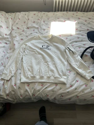 Vit sweatshirt från C.P. Company - Vit sweatshirt från C.P. Company med svart logga på bröstet och en ikonisk linsdetalj på ärmen. Tröjan har rund halsringning och långa ärmar. Perfekt för en avslappnad och trendig stil.
