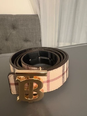 Rutig Burberry-bälte med guldspänne - Snyggt bälte från Burberry med klassiskt rutigt mönster i beige, rosa och svart. Bältet har ett stort guldigt spänne med Burberrys ikoniska TB-logga. Perfekt accessoar för att lyfta vilken outfit som helst. 130cm