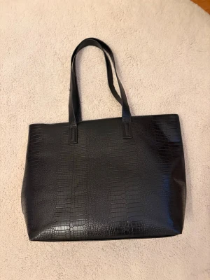 Svart shoppingväska i crocopräglat skinn - Stilren svart shoppingväska med crocopräglat mönster och långa axelband. Väskan är rymlig och har en klassisk rektangulär form. Perfekt för att bära med sig allt man behöver under dagen. Materialet är konstläder med glansig finish.