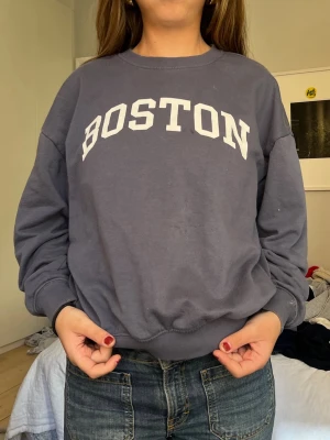Grå sweatshirt med BOSTON-tryck - Cool grå sweatshirt med vit BOSTON-text på bröstet. Klassisk rund halsringning och ribbade muddar vid ärmar och midja. Perfekt för en avslappnad streetstyle-look.