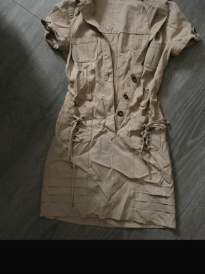 Beige kort klänning med snörning Märke BEBE sommars/ halloween - KÖPT FRÅN USA STORLKE 0 Snygg beige kortklänning med knappar framtill, snörningsdetaljer vid fickorna och korta ärmar. Klänningen har en figurnära passform och flera fickor framtill. Perfekt för dig som gillar en stilren och trendig look. Knapparna är däremot lösa men som går att sy fast igen. Gula fläckar men som också går nog att få bort i tvätten 