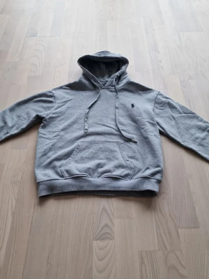 Grå hoodie från Polo Ralph Lauren - Klassisk grå hoodie från Polo Ralph Lauren med broderad logga på bröstet. Hoodien har huva med dragsko, magficka och ribbade muddar vid ärmslut och nederkant. Perfekt för en avslappnad och stilren look.