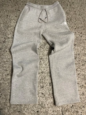 Grå mjukisbyxor från The North Face - Snygga grå mjukisbyxor från The North Face med dragsko i midjan och två sidofickor. Byxorna har en avslappnad passform och är tillverkade i mjukt material som är perfekt för chill dagar. Loggan syns på vänster lår.