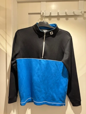 Tröja från FootJoy | JR XL - Snygg långärmad sporttröja från FootJoy i svart och blått med halv dragkedja framtill. Tröjan har hög krage med FJ-logga och kontrastsömmar. Perfekt för golf eller annan träning. Materialet är mjukt och stretchigt för bra rörelsefrihet.