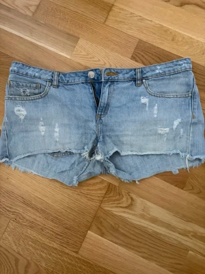 Ljusblå slitna jeansshorts - Säljer ett par ljusblå jeansshorts med slitna detaljer och fransig kant. Klassisk femficksmodell med bälteshällor och knappgylf. Perfekta för sommaren och ger en avslappnad vibe.