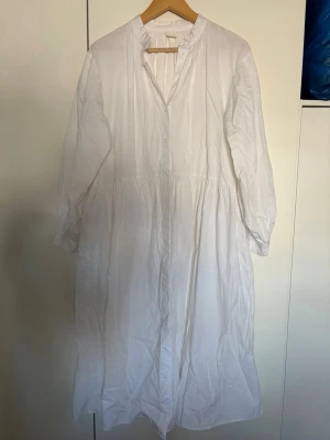 Vit långklänning från H&M i bomull - Luftig och stilren vit långklänning från H&M i mjuk bomull. Klänningen har lång ärm, v-ringning och knappar framtill. Perfekt för en fräsch och enkel look under varmare dagar. Har fickor på sidan 