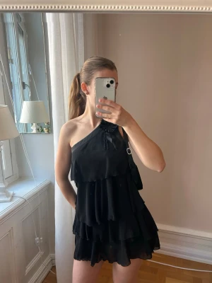 Svart one shoulder volangklänning - Svart kortklänning med one shoulder-design och flera lager volanger. Dekorativ blomdetalj på axeln och är tillverkad i ett lätt. Ger snygg siluett. Trendig och perfekt till festligheter✨