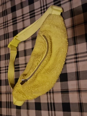 Gul fluffig magväska med dragkedja - Säljer en cool gul magväska i mjukt teddy-material med justerbart band och silverfärgad dragkedja framtill. Perfekt accessoar för att sticka ut och ha med det viktigaste på språng.