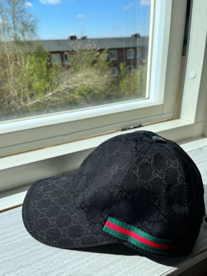 Svart Gucci keps med GG-mönster - Snygg svart keps från Gucci med klassiskt GG-monogram över hela kepsen. På sidan finns en grön och röd rand som ger en lyxig touch. Justerbar rem baktill för perfekt passform. Materialet är canvas med exklusiv känsla.