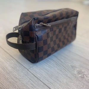 Brun och svart rutig necessär - Snygg necessär med klassiskt brunt och svart rutmönster. Den har en praktisk dragkedja och ett handtag på sidan för enkel transport. Perfekt för att hålla ordning på dina saker.