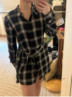 Svartvit rutig skjortklänning med bälte - Svartvit rutig skjortklänning med lång ärm och knytbälte i midjan. Klassisk krage och avslappnad passform, perfekt för en trendig look. Klänningen är kort och har ett stilrent rutmönster.