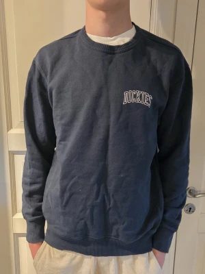 Mörkblå sweatshirt från Dickies - Mörkblå sweatshirt från Dickies med rund halsringning och vit logotyp på bröstet. Tröjan har ribbade muddar vid ärmslut och nederkant. Perfekt för en avslappnad och stilren look.
