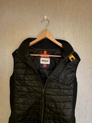 Svart quiltad dunväst Parajumpers - Snygg svart dunväst från Parajumpers med quiltad design och dragkedja framtill. Västen har en klassisk ståkrage och en gul detalj vid axeln. Perfekt för lager på lager under kyliga dagar. Storleken är 176 och fråga om ni vill se bild 