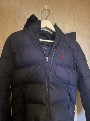 Mörkblå pufferjacka Polo Ralph Lauren - Snygg mörkblå pufferjacka från Polo Ralph Lauren med huva och dragkedja framtill. Jackan har två sidofickor med dragkedja och den klassiska röda Polo-loggan på bröstet. Perfekt för kalla vinterdagar och riktigt skön att bära. Det finns en defekt men den syns inte långt ifrån passa på att köpa i förväg till vintern för en jätte snygg jacka 