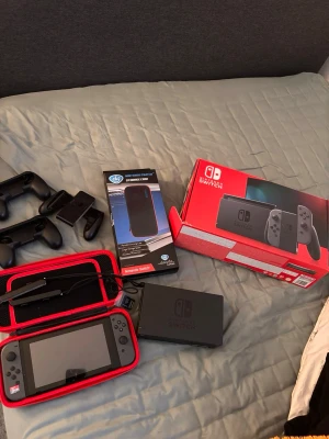 Nintendo Switch med tillbehör och väska - Nintendo Switch spelkonsol i grått med originalkartong, två Joy-Con-kontroller, svart dockningsstation, laddare, handkontrollshållare och en röd bärväska. Perfekt för gaming hemma eller på språng. Inkluderar även ett extra tillbehörspaket.