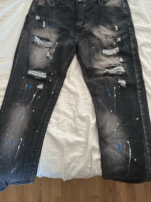Svarta distressed jeans med färgstänk - Säljer ett par svarta jeans med slitna detaljer och hål på framsidan. Jeansen har blå och vita färgstänk både fram och bak. Rak passform