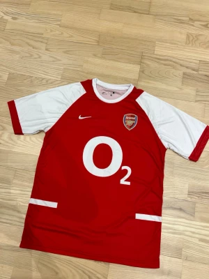 Arsenal Nike matchtröja röd/vit - Klassisk Arsenal matchtröja från Nike i rött med vita ärmar och detaljer. Stort O2-tryck på bröstet och Arsenal-logga på vänster sida. Perfekt för fotbollsfans som vill visa sitt stöd för laget. Tillverkad i lätt och ventilerande material.