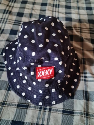 Mörkblå bucket hat med vita prickar - Snygg mörkblå bucket hat från XX-XY med vita prickar och en röd patch framtill. Hatten är i mjukt tyg och har klassisk form med nedvikt brätte. Perfekt accessoar för en cool och avslappnad stil.