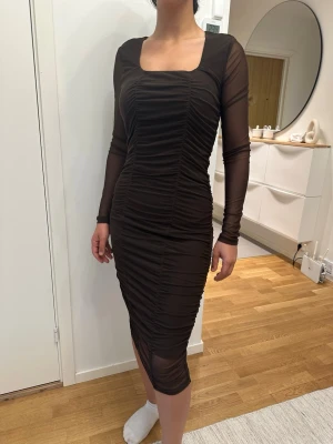 Brun långärmad klänning från BikBok - Snygg brun långklänning från BikBok med långa ärmar i mesh och rynkade detaljer längs hela klänningen. Fyrkantig ringning och figurnära passform ger en trendig look. Perfekt för dig som vill sticka ut med stil.