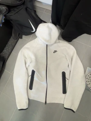 Nike Tech Fleece hoodie ljusgrå - Nike Tech Fleece hoodie i ljusgrått med svarta detaljer. Dragkedja framtill, två svarta fickor med dragkedja och Nike-logga på bröstet. Mjuk och sportig design, perfekt för träning eller chill. Tillverkad i bomullsblandning.