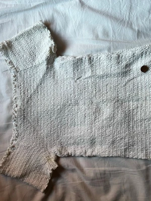 Vit ribbad topp med volangkant - Supersöt vit ribbad topp med korta ärmar och volangkant längs ärmslut och nederkant. Toppen är stretchig och har en smickrande passform som sitter tajt mot kroppen. Perfekt att styla med jeans eller kjol för en trendig look.