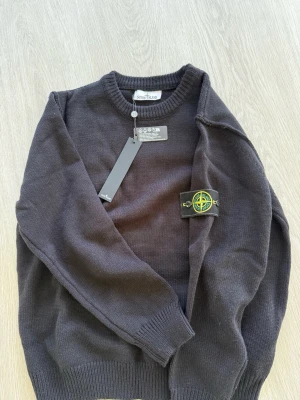 Svart stickad tröja Stone Island - Säljer en svart stickad tröja från Stone Island med klassisk rund hals och ribbade muddar. Tröjan har den ikoniska Stone Island-loggan på vänster ärm och är tillverkad i mjukt material. Perfekt för dig som gillar streetwear och stilrena plagg.