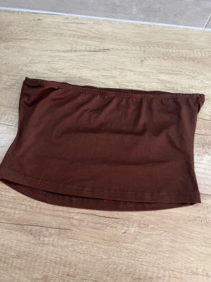 Brun bandeau tubtopp - En enkel och stilren brun bandeau tubtopp i mjukt material. Toppen är axelbandslös och har en elastisk kant upptill för att sitta på plats. Perfekt att matcha med höga jeans eller kjol.