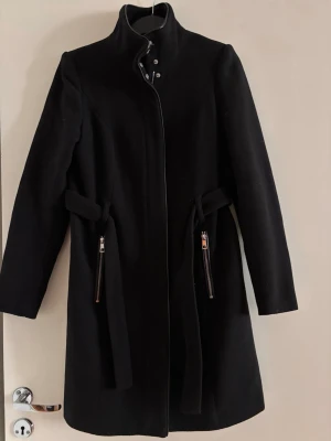 Svart kappa från Vero Moda - Stilren svart kappa från Vero Moda med hög krage, dragkedjor vid fickorna och knytskärp i midjan. Klassisk rak modell som passar perfekt till kyliga dagar. Materialet är mjukt och ger en snygg siluett.