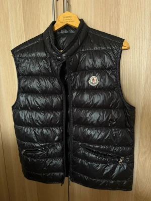 Svart Moncler väst - Snygg svart dunväst från Moncler med quiltad design och hög krage. Västen har två dragkedjefickor framtill och Monclers klassiska logga på bröstet. Perfekt för lager-på-lager och ger en stilren look. Materialet är glansigt och lätt. Storlek: 2 (S/M)