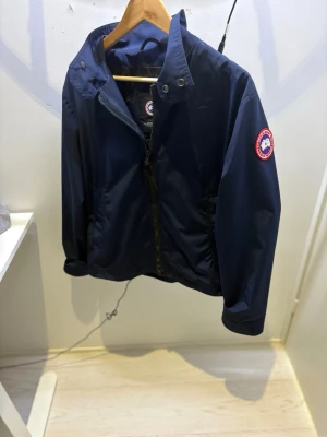  Canada Goose rosedale - Snygg mörkblå vindjacka från Canada Goose med dragkedja framtill, hög krage och logotyp på vänster ärm. Jackan har två sidofickor och reflekterande detalj i nacken. Perfekt för blåsiga dagar och enkel att matcha. Brand new never used