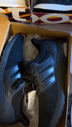 Svarta Adidas Ultraboost sneakers - Snygga helsvarta Adidas Ultraboost sneakers med stickad ovandel och klassiska tre ränder på sidan. Skorna har en chunky Boost-sula för maxad komfort och sportig look. Perfekta för dig som gillar stilrena och bekväma sneakers med modern känsla.