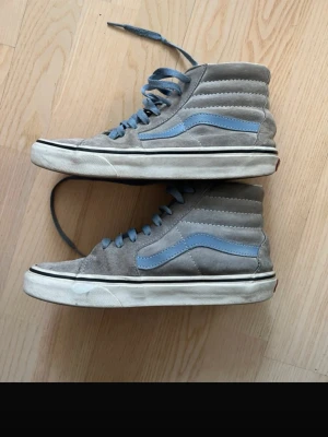 Gråa Vans med blå detaljer - Gråa Vans Sk8-Hi sneakers i mocka med ljusblå sidestripe och matchande blå skosnören. Klassisk hög modell med vit sula och brun yttersula. Perfekta för en avslappnad streetstil.