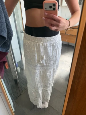 Vit långkjol med broderade detaljer - Superfin vit långkjol med broderade blommor och spetsdetaljer. Kjolen har resår i midjan och är tillverkad i ett lätt bomullstyg, perfekt för varma dagar. Den har en romantisk och bohemisk känsla med vackra mönster och volanger. STÅR ATT DEN ÄR XL/L MEN PASSAR SOM EN XS/S!!!