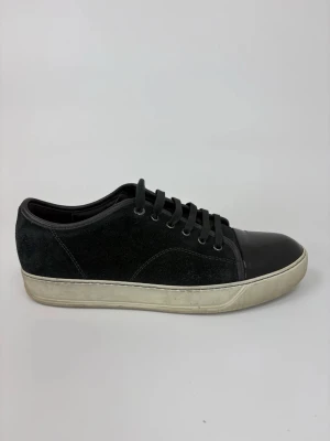 Svarta Lanvin sneakers med lackad tå - Stilrena svarta sneakers med ovandel i mocka och glansig, lackad tå. Skorna har svarta skosnören och en vit sula som ger en klassisk kontrast. Perfekta för dig som gillar en clean och modern look med lite extra edge.