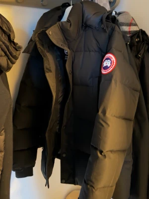 Svart dunjacka från Canada Goose - Säljer en svart dunjacka från Canada Goose med klassisk patch på ärmen. Jackan har hög krage, dragkedja och tryckknappar framtill samt stora quiltade partier för extra värme. Perfekt för kalla vinterdagar och riktigt snygg streetstil.