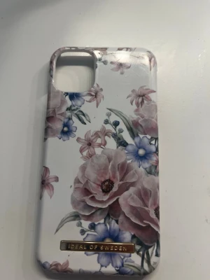 Blommigt mobilskal i rosa och blått iPhone 11 - Snyggt mobilskal från iDeal of Sweden med blommigt mönster i rosa, blått och grönt på vit bakgrund. Skalets form är anpassad för en mobil med dubbla kameror. Tillverkat i hårdplast med en metallbricka med logga längst ner.