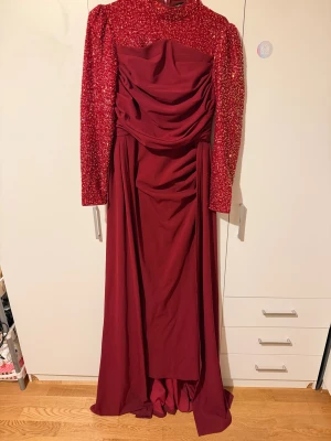 Röd långklänning med paljetter - Säljer röd långklänning med glittriga paljetter på överdelen och ärmarna. Klänningen har draperad front, hög hals och långa ärmar. Kjolen är mjukt fallande och ger en riktigt elegant look. Perfekt för dig som vill sticka ut på bal,fest eller klänning. Andvänt en gång! Köpt för 700kr