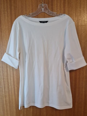 Vit topp från Ralph Lauren - Klassisk vit topp från Ralph Lauren med båthals och uppvikta ärmar. Tillverkad i 94% mjuk bomull och 6% elastan som känns skön mot huden. Enkel och stilren design med sprund i sidorna och en diskret broderad logga på ena sidan (se bild). Aldrig använd men inga taggar kvar.