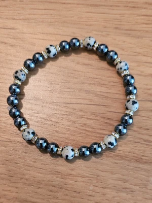 Armband med svarta och vita pärlor - Snyggt armband med halvädelstenar som Dalmatinerjaspis och Hematit. Elastiskt band gör det enkelt att ta på och av. Perfekt accessoar för att lyfta din stil. Det levereras i en söt liten påse tillsammans med information om kristallernas magiska egenskaper. 