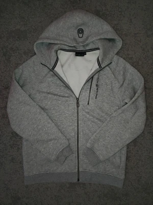 Grå hoodie från Sail Racing - Snygg grå hoodie från Sail Racing med dragkedja framtill och en extra bröstficka med dragkedja. Broderad logga på bröstet och huva med svartvitt kantband. Mjuk insida och ribbade muddar vid ärmslut och nederkant.
