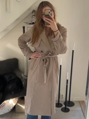 Beige trenchcoat med knytskärp - Stilren beige trenchcoat med klassisk dubbelknäppning och brett slag. Knytskärp i midjan och svarta knappar framtill. Lång modell som passar perfekt till jeans eller klänning. Materialet ser ut att vara bomullsblandning.
