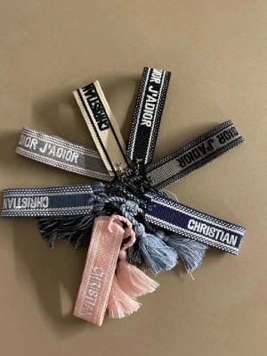 Christian Dior broderade armband - Broderade armband från Christian Dior i olika färger som svart, blå, rosa och beige. Armbanden har texten 'Christian Dior' eller 'J'Adior' och är dekorerade med tofsar i matchande färg. Perfekt för att ge en cool och exklusiv touch till din stil.