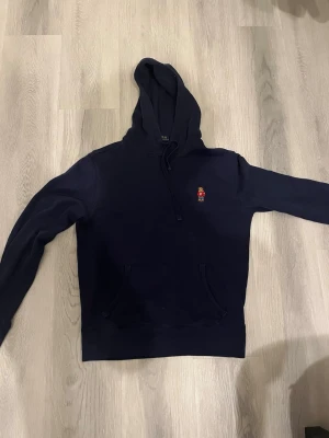 Mörkblå hoodie från Polo Ralph Lauren - Snygg mörkblå hoodie från Polo Ralph Lauren med broderad björn på bröstet. Klassisk känguruficka framtill och justerbar huva med dragsko. Perfekt för en chill och stilren look. Mjukt material som känns skönt mot huden.
