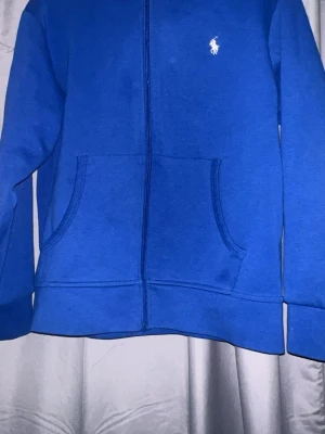 Blå hoodie från Polo Ralph Lauren - Blå hoodie med dragkedja från Polo Ralph Lauren. Klassisk design med fickor framtill och vit broderad logga på bröstet. Tillverkad i mjukt bomullsmaterial och har en skön huva. Perfekt för en avslappnad och stilren look.