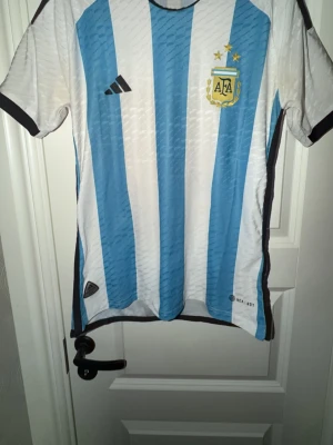 Argentina fotbollströja Adidas - Säljer en klassisk Argentina fotbollströja från Adidas med blå och vita vertikala ränder, svarta detaljer och AFA-emblem med tre stjärnor på bröstet. Tröjan är kortärmad och tillverkad i ett lätt och ventilerande material, perfekt för fotboll eller träning.