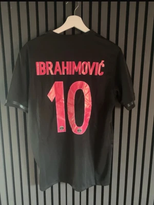Svart PSG tröja Ibrahimović #10 - Säljer en svart Paris Saint-Germain matchtröja från Nike med Ibrahimović och nummer 10 på ryggen. Tröjan har vita detaljer, PSG-logga och Fly Emirates-tryck på bröstet. Perfekt för fotbollsfans som vill sticka ut på läktaren eller planen.